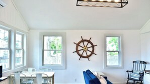 Un salón luminoso y amplio con un gran timón de barco decorativo en la pared, un sofá azul y una zona de comedor con una mesa y sillas blancas.