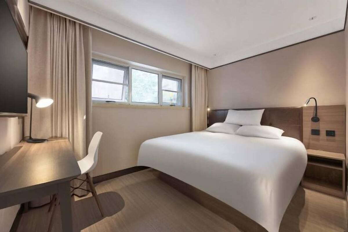 A hotel room with a bed, a desk, a chair, a window, and a lamp.