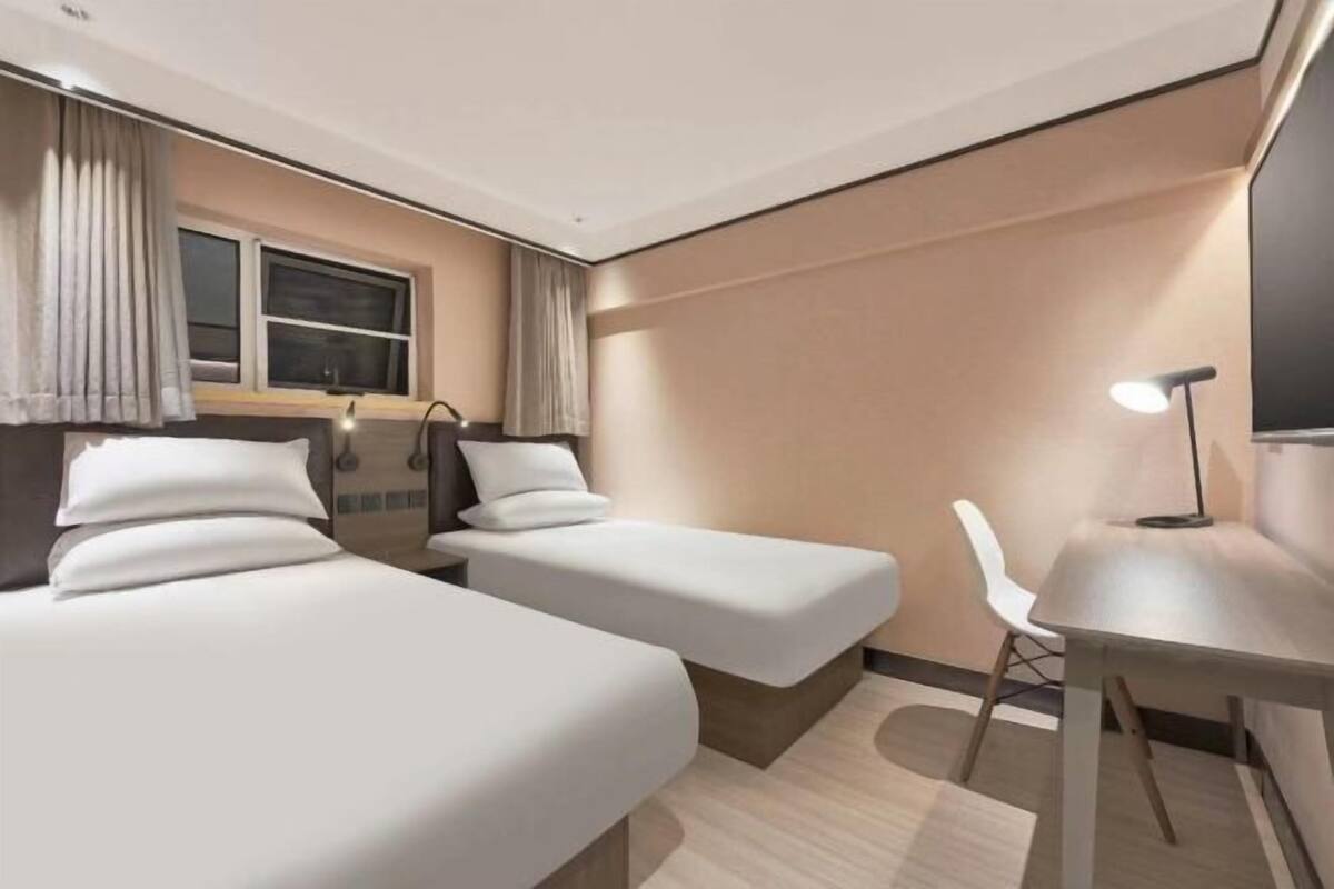 A hotel room with two beds, a desk, a chair, a lamp, and a window with curtains.