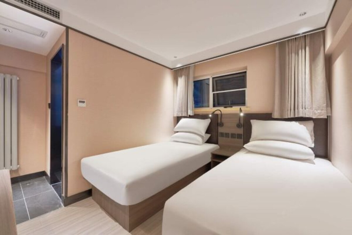 Habitación de hotel con dos camas individuales, una ventana con cortinas, un radiador y una puerta que da a otra habitación.