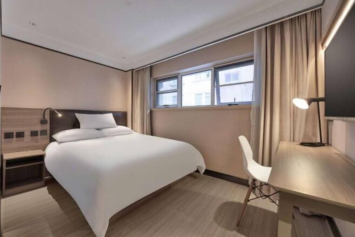 A hotel room with a bed, bedside table, lamp, chair, and a window with curtains.