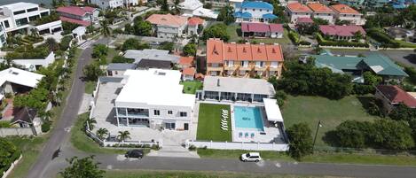 An aerial view of a coastal residential area with a large building featuring a swimming pool, surrounded by houses with red roofs and a marina with boats.
