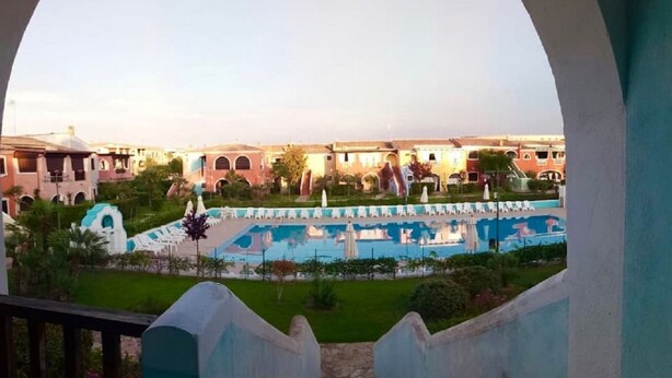 A pool area with multiple lounge chairs and a central water feature, surrounded by buildings with arched windows and a well-maintained lawn. - BORGO SAN BASILIO: MOONLIGHT HOUSE - CON PISCINA E ANIMAZIONE DIURNA E SERALE (Pisticci)