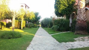 A stone pathway leading through a well-maintained garden with various plants and trees, flanked by buildings with arches and a clock tower. - BORGO SAN BASILIO: MOONLIGHT HOUSE - CON PISCINA E ANIMAZIONE DIURNA E SERALE (Pisticci)