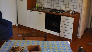 Une cuisine compacte avec des meubles blancs, une cuisinière à gaz et un lave-vaisselle. Une table bleue recouverte d’une nappe à carreaux se trouve au premier plan.