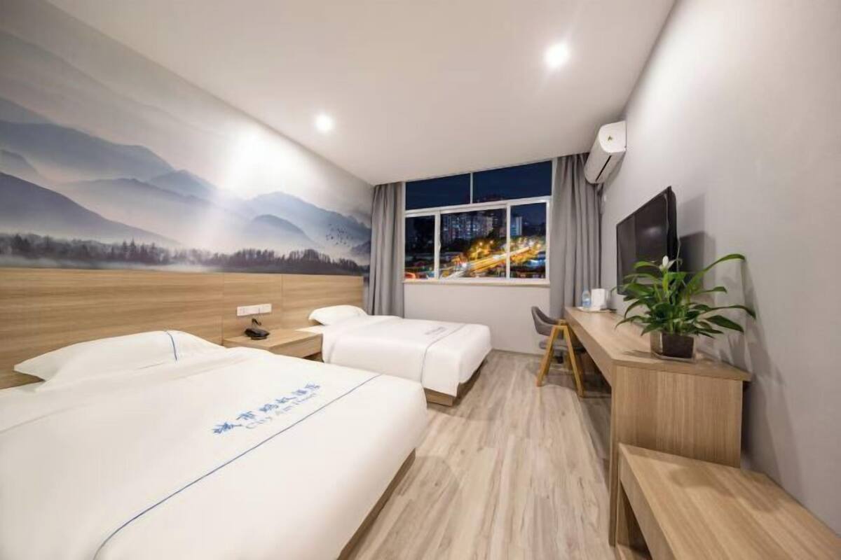 A hotel room with two beds, a desk, a TV, and a window with a city view.