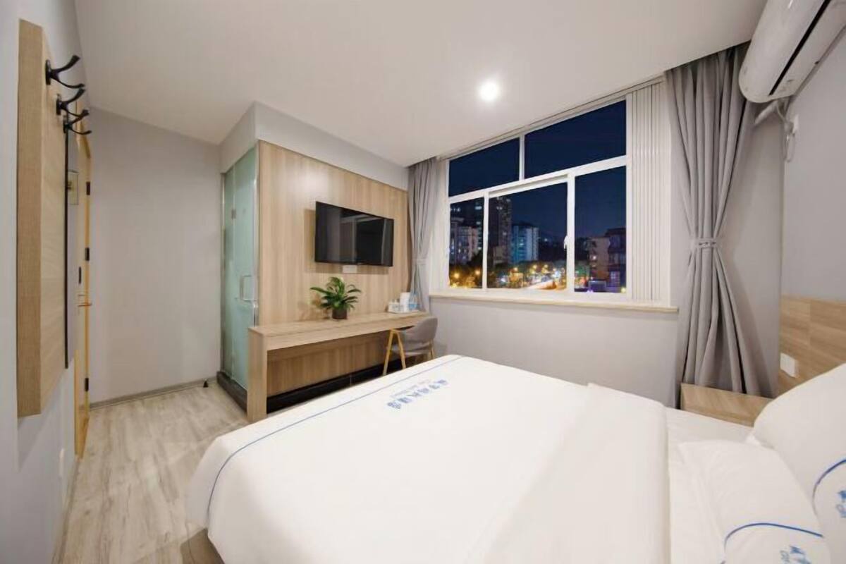 A hotel room with a bed, a TV mounted on the wall, a desk with a chair, and a window with a city view.