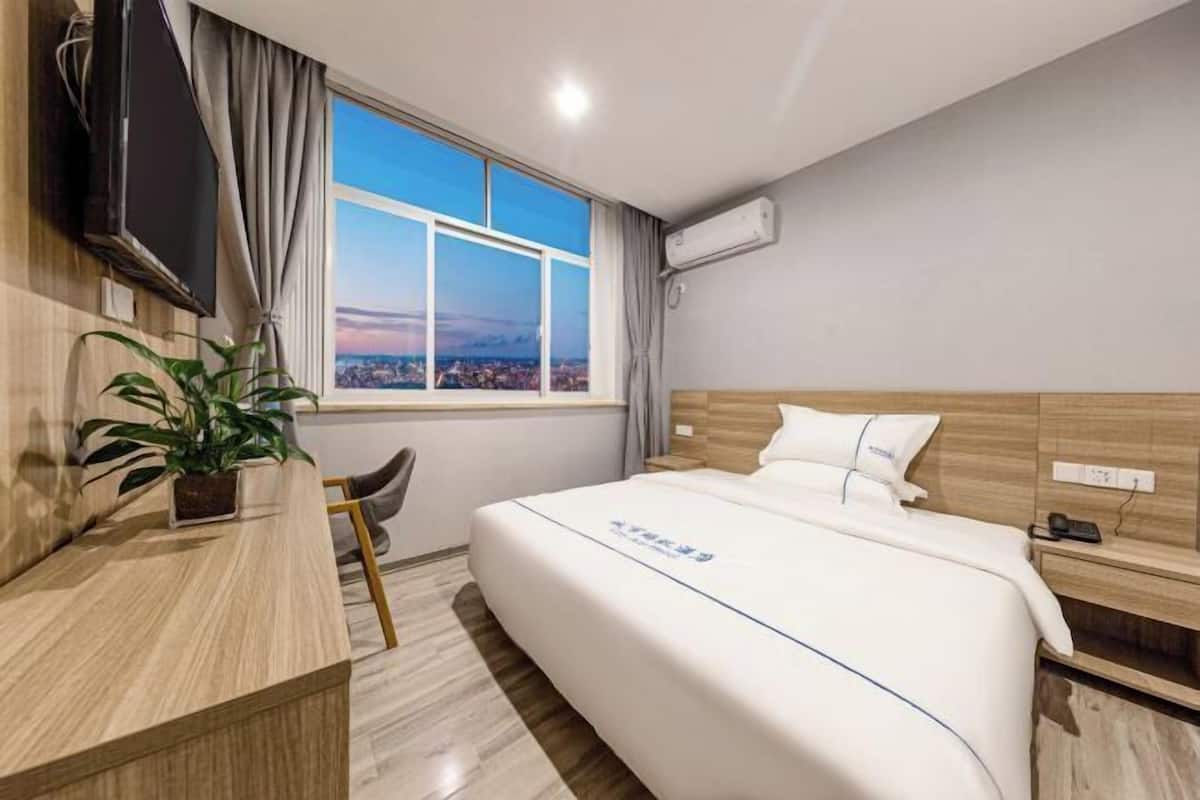 A hotel room with a bed, a desk, a TV, a window with a city view, and an air conditioning unit.