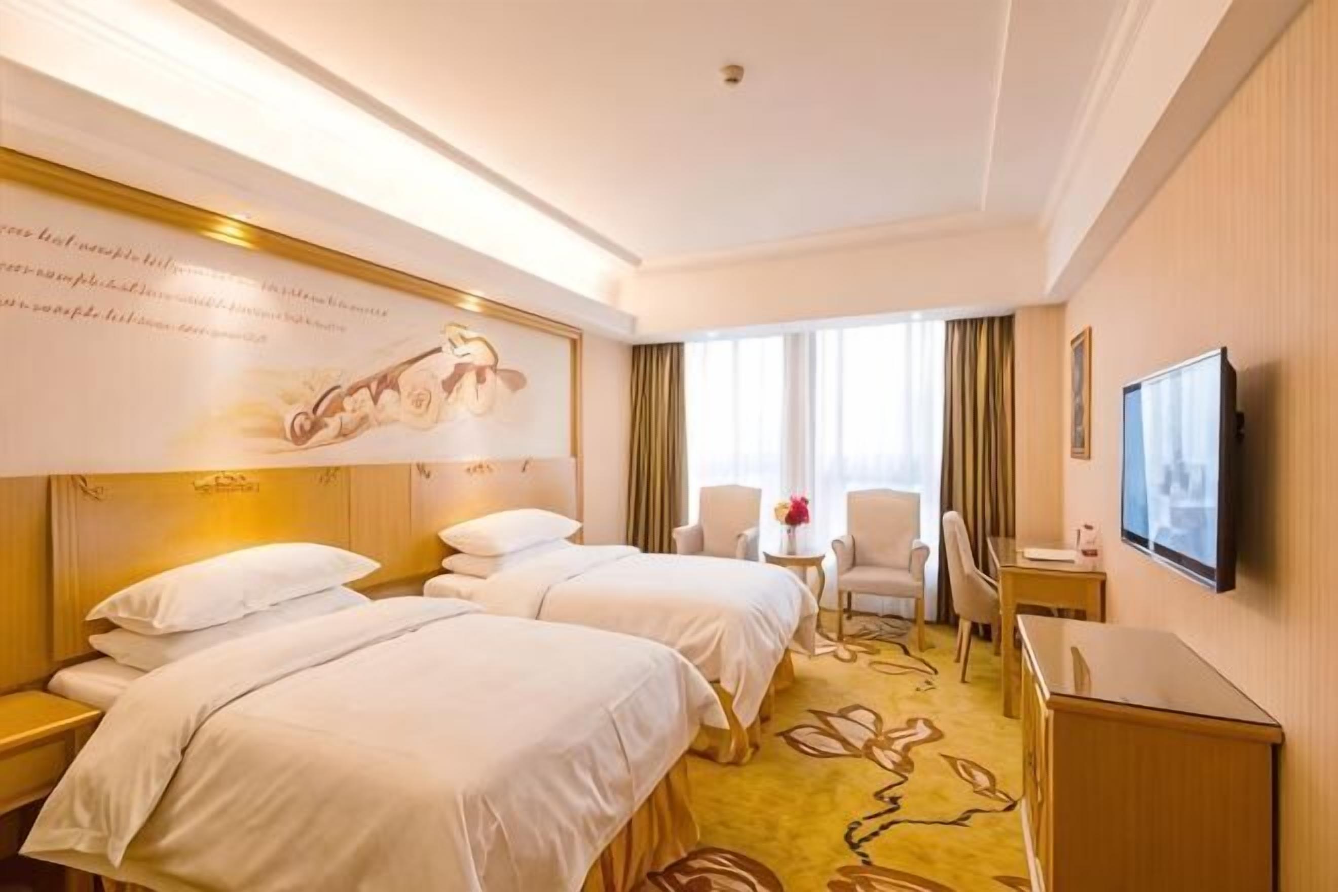 A hotel room with two beds, a desk, a chair, a TV, and a window with curtains.