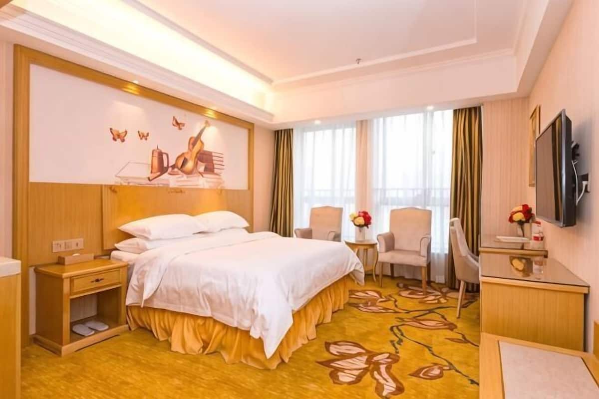 A hotel room with a bed, bedside tables, a desk, a chair, a TV, and a window with curtains.