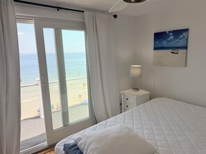 A bedroom with a bed, a nightstand, a lamp, a painting, and a view of the beach. - Wake up to spectacular sunrises and the sound of waves at this great beach house (Marshfield)