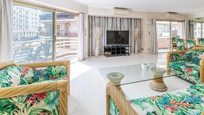 Un salón amplio con balcón, amoblado con sillas de mimbre y un televisor de pantalla plana.