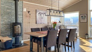 A modern dining area with a wooden table, grey upholstered chairs, a stone wall, a fireplace, and a city view through large windows.