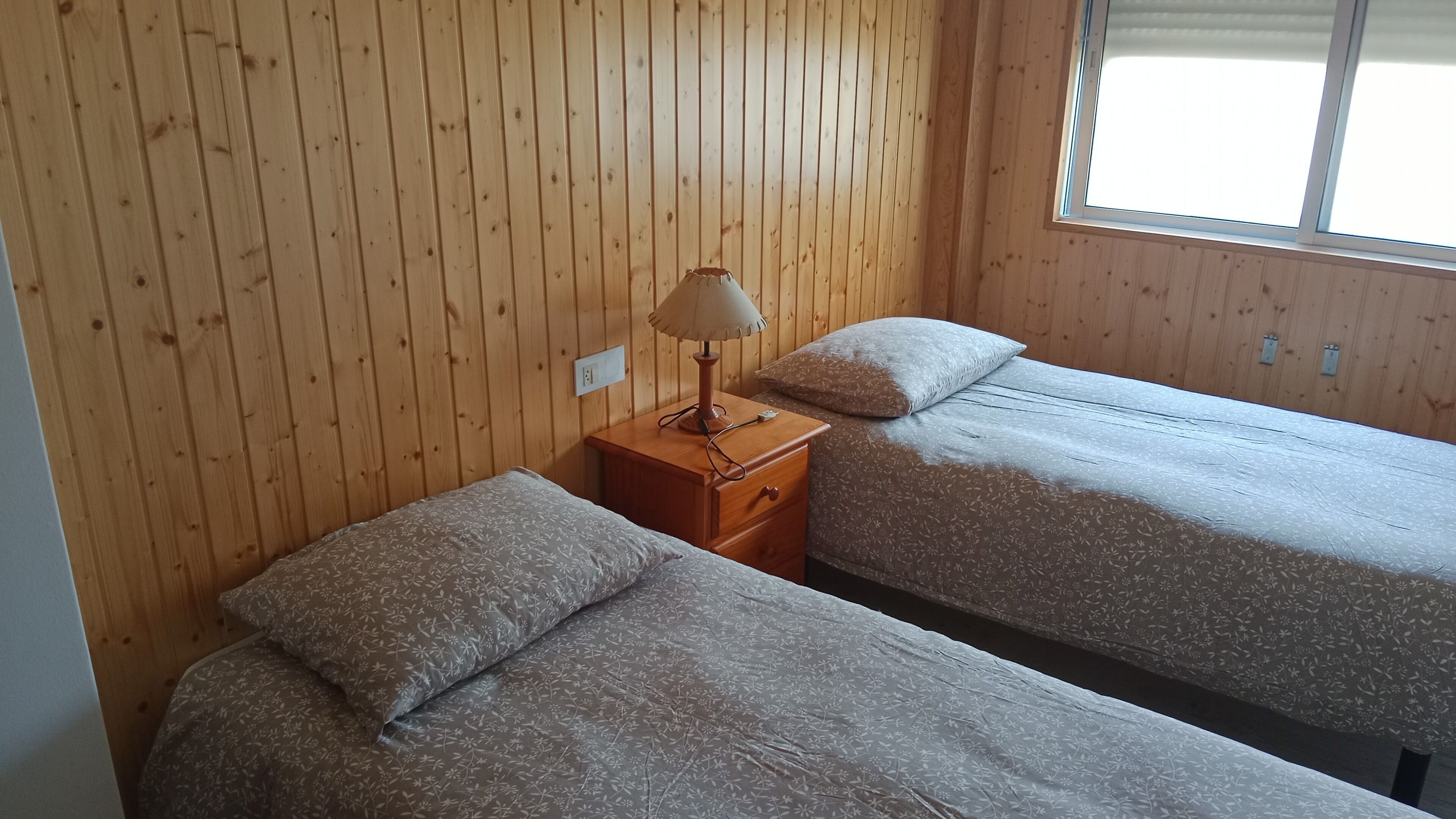 A room with wooden panel walls, two beds with floral-patterned bedding, a nightstand with a lamp, and a window with blinds.