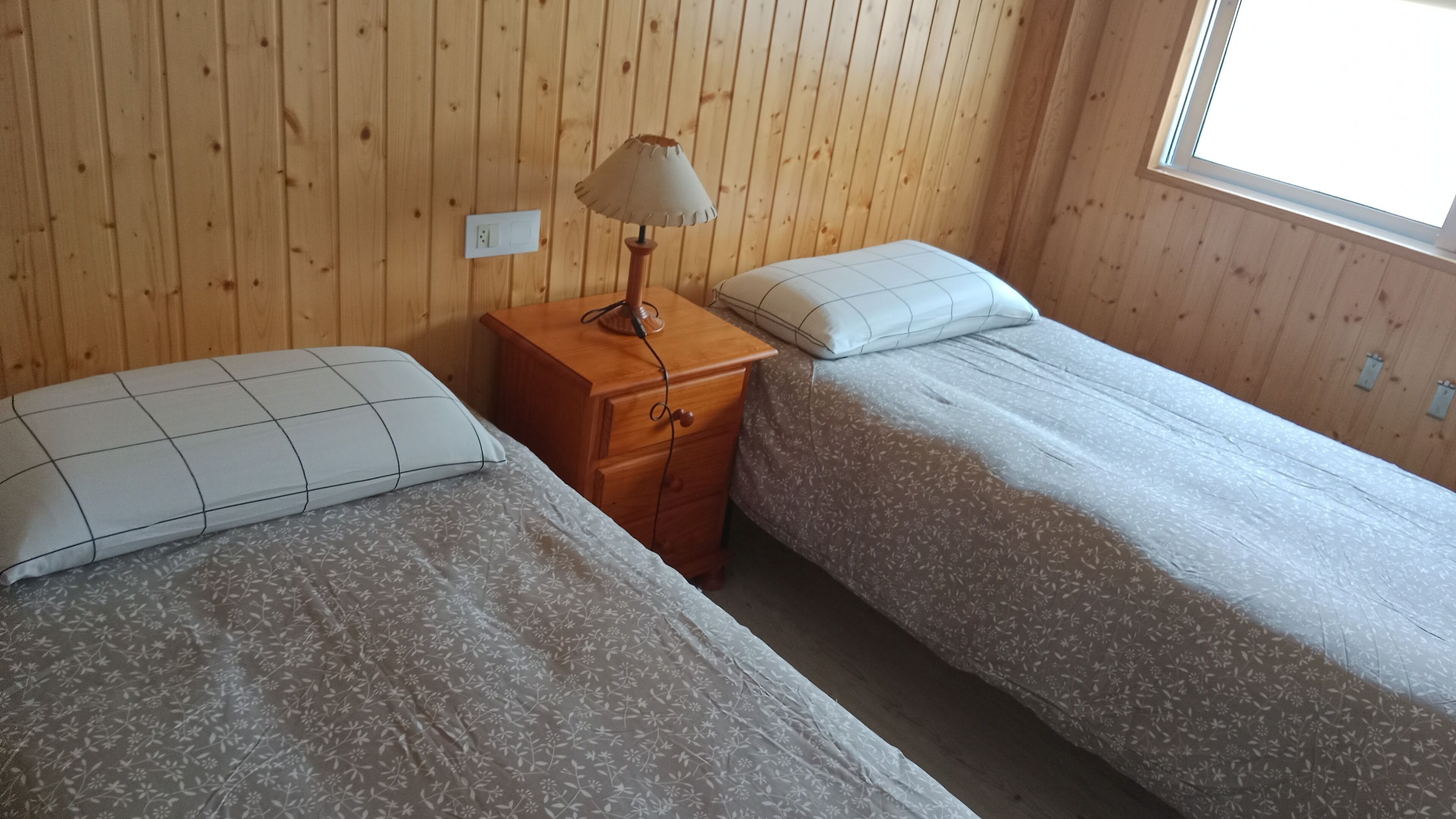 Quarto com duas camas de solteiro, uma mesa de cabeceira de madeira com abajur e uma janela.