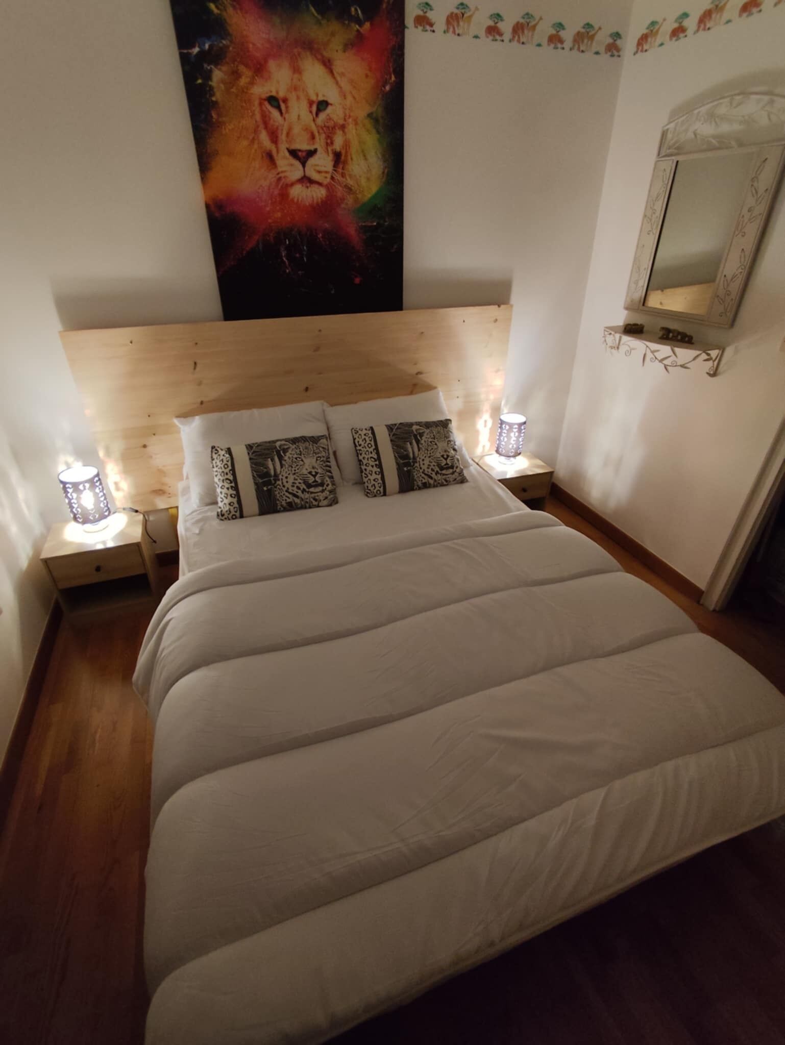 Une chambre à coucher avec une tête de lit en bois, un grand lit avec une literie blanche, des coussins à motifs léopard, deux lampes de chevet et un miroir fixé au mur.