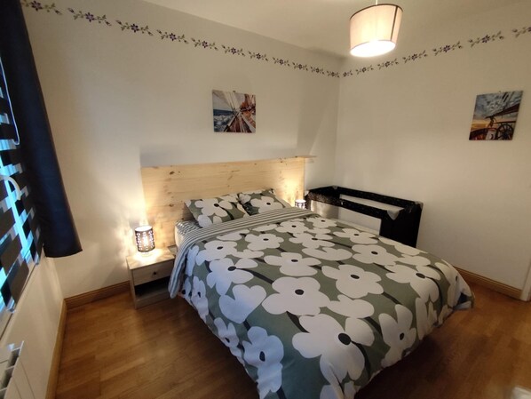 A bedroom with a wooden headboard, a floral bedspread, a nightstand with a lamp, and two wall-mounted pictures.