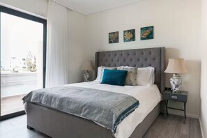 Une chambre d’hôtel moderne avec un grand lit, une tête de lit grise et une vue sur la ville par une grande fenêtre.