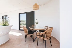 Un espace repas extérieur moderne, avec des chaises en osier et une table en verre, un grand pot de fleurs blanc et une vue à travers une porte-fenêtre coulissante.