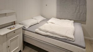 A single bed with a grey mattress and white bedding, a bedside table with a book, and a window with blinds. - 1 bedroom nice apartment in Kvås (Kvås)