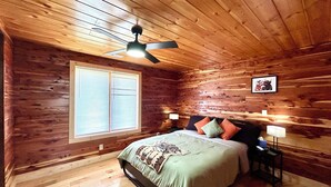 Un dormitorio con paneles de madera, una cama, un ventilador de techo, una ventana con persianas y una mesita de noche con lámpara.