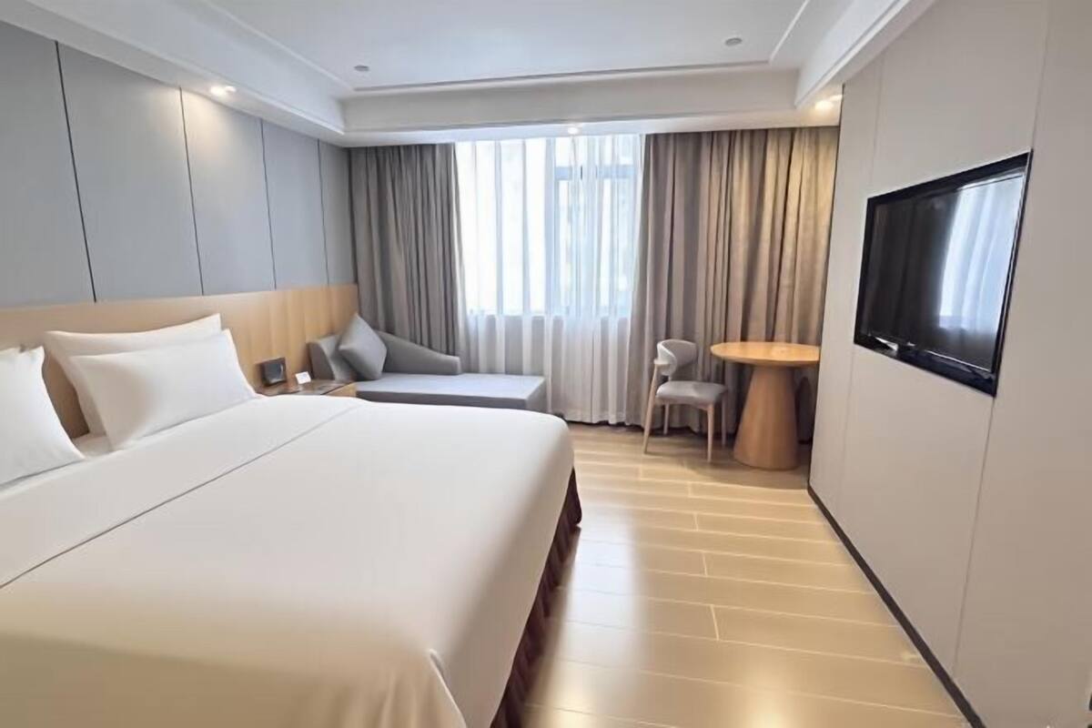 A hotel room with a large bed, a seating area with a table and chairs, a TV mounted on the wall, and a window with curtains.