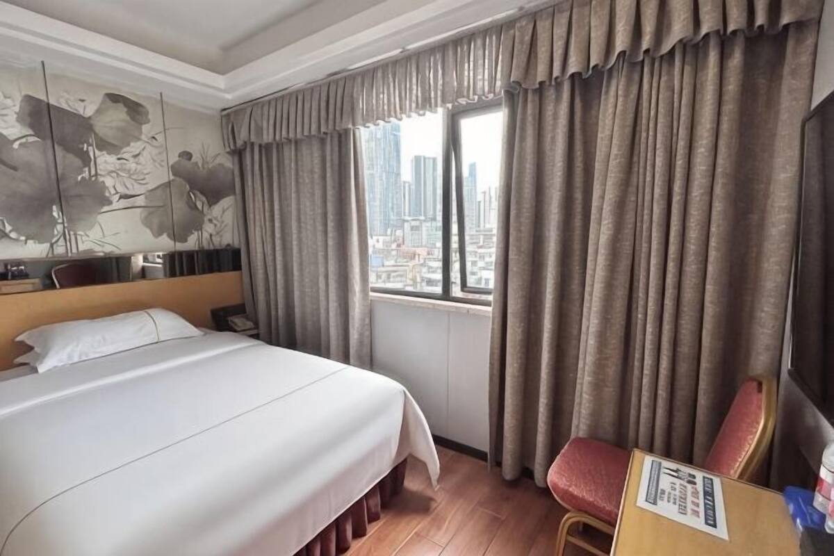A hotel room with a bed, a chair, a window with curtains, and a city view.