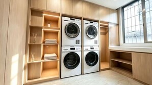 A laundry room with two stacked front-loading washing machines, wooden shelving units, and a large window.