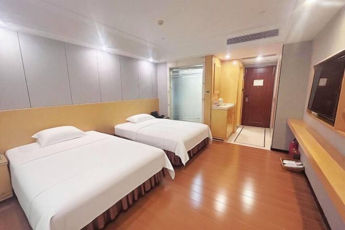 A hotel room with two beds, a wooden floor, a TV, and a bathroom with a mirror and sink.