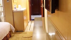 A hotel hallway with a bed, a small desk with a mirror, and a door at the end.