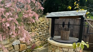Una chimenea de piedra al aire libre con techo metálico, rodeada por un muro de piedra y arbustos en flor.