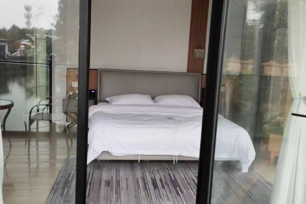 A bedroom with a bed, a glass partition, and a view of the outdoors.