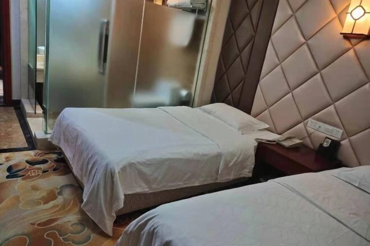 A hotel room with two beds, a glass partition, a wall-mounted light, and a patterned carpet.