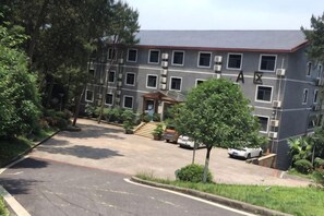 Un edificio de varios pisos con estacionamiento, árboles y un pequeño pabellón de entrada.