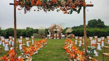 Espaço para casamento ao ar livre, com arco de flores, cadeiras dispostas em fileiras e um painel decorativo.