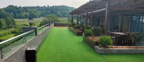 A wooden deck with a green artificial turf lawn, a pergola structure, and a view of a forested area.