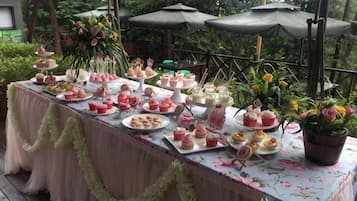 An outdoor table with various food items and drinks, surrounded by greenery and umbrellas.