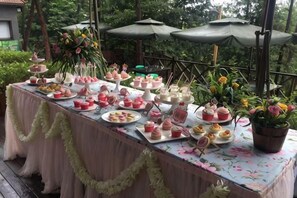 Une table installée à l’extérieur, sur laquelle on trouve divers aliments et boissons, le tout entouré de verdure et d’ombrelles.