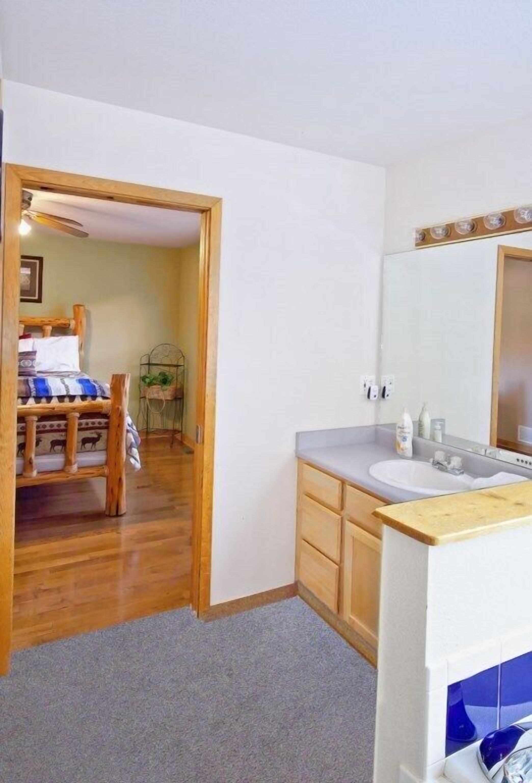 A bedroom with a wooden bed, a bedside table, a small desk, and a chair. There is a bathroom with a sink, mirror, and toiletries.