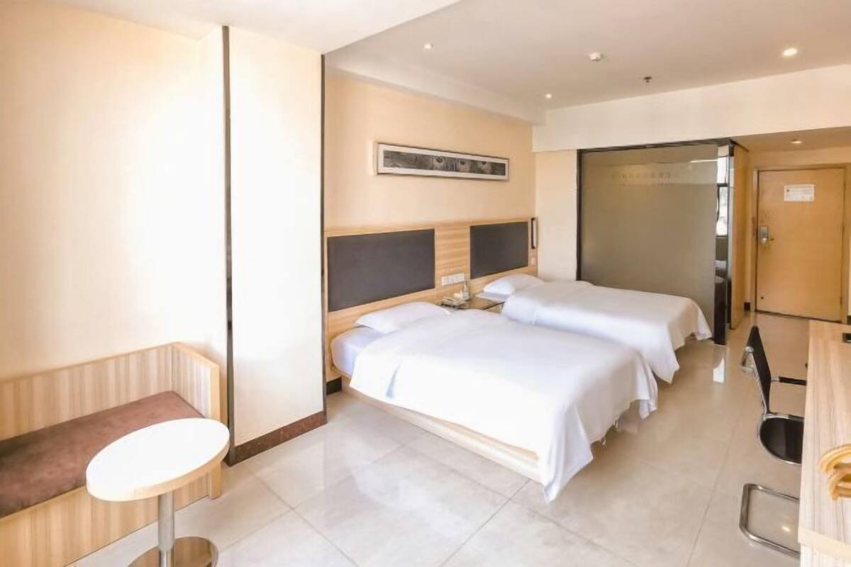 A hotel room with two beds, a small round table, a bench, and a door leading to a bathroom.