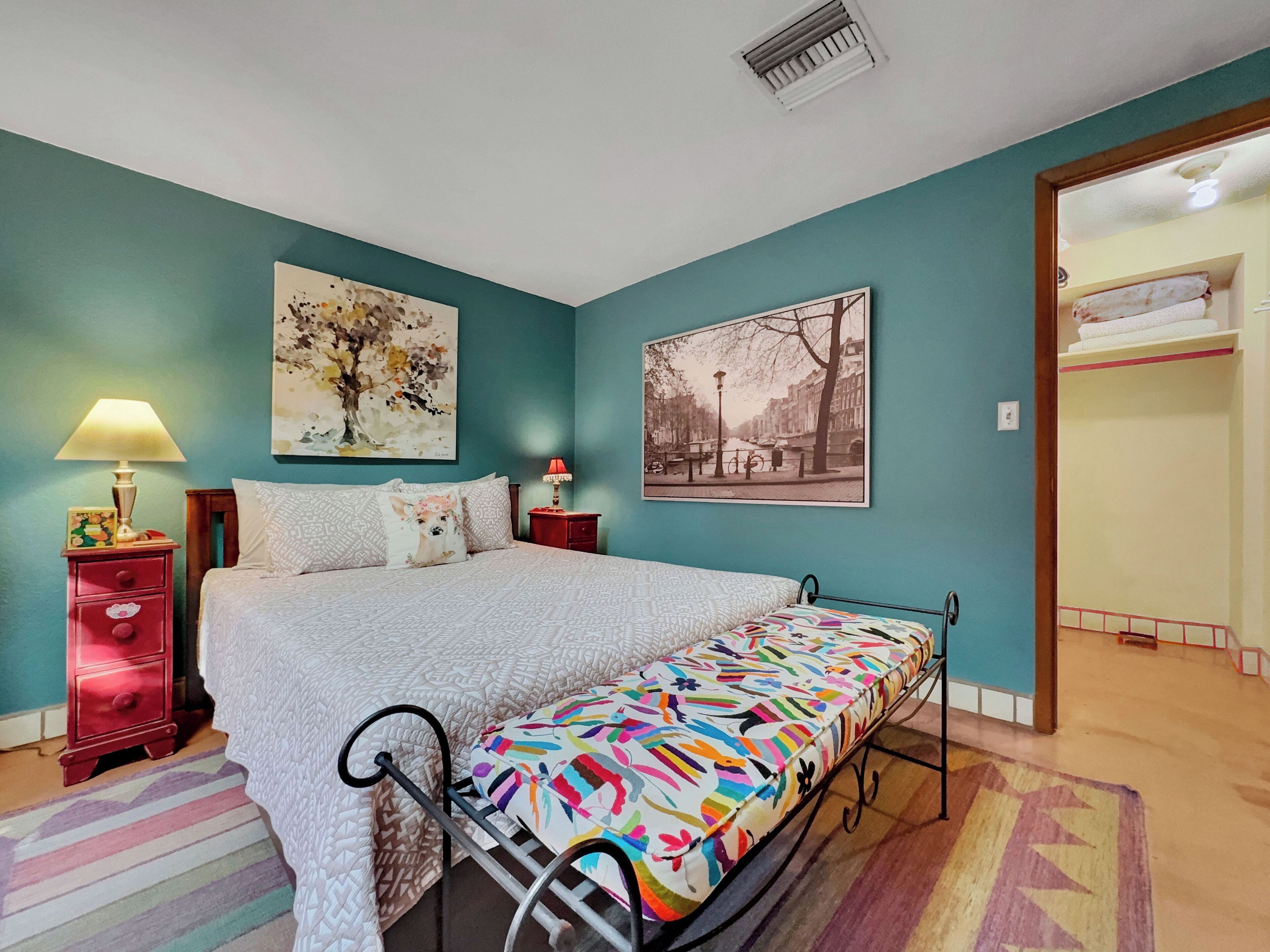 Une chambre à coucher comprenant un lit, deux tables de chevet, une commode rouge, un matelas coloré et deux œuvres d’art murales.