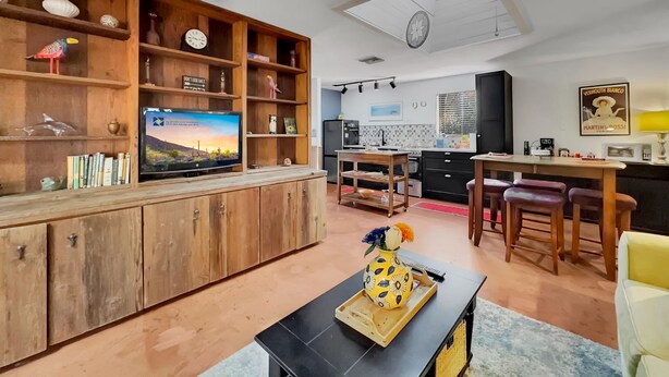 Un salon avec un meuble de rangement en bois pour l’équipement audiovisuel, un téléviseur à écran plat, un coin repas avec une table et des chaises, et une cuisine à l’arrière-plan.