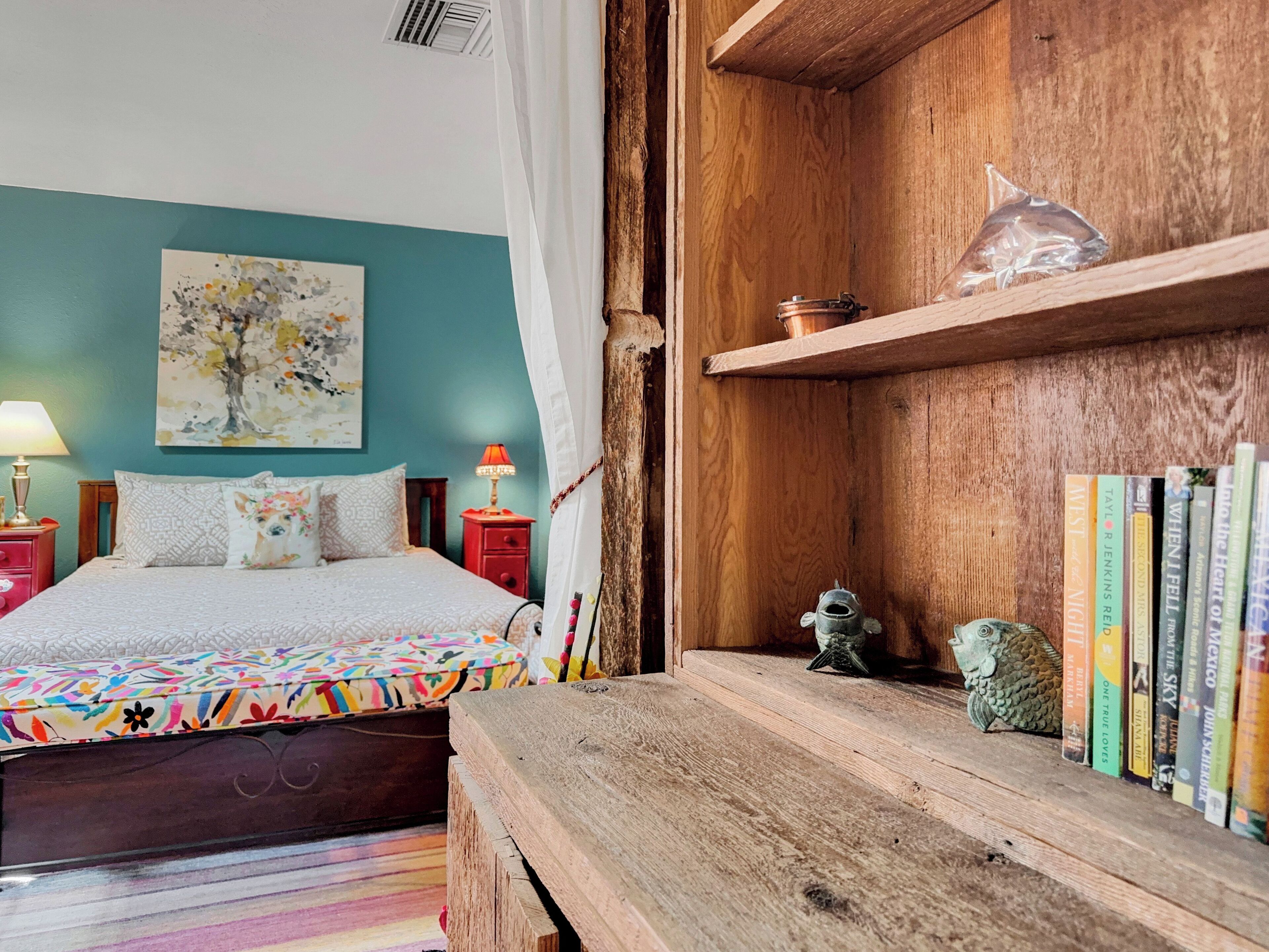 A bedroom with a bed, a nightstand, a lamp, a bookshelf, and a wall painting.