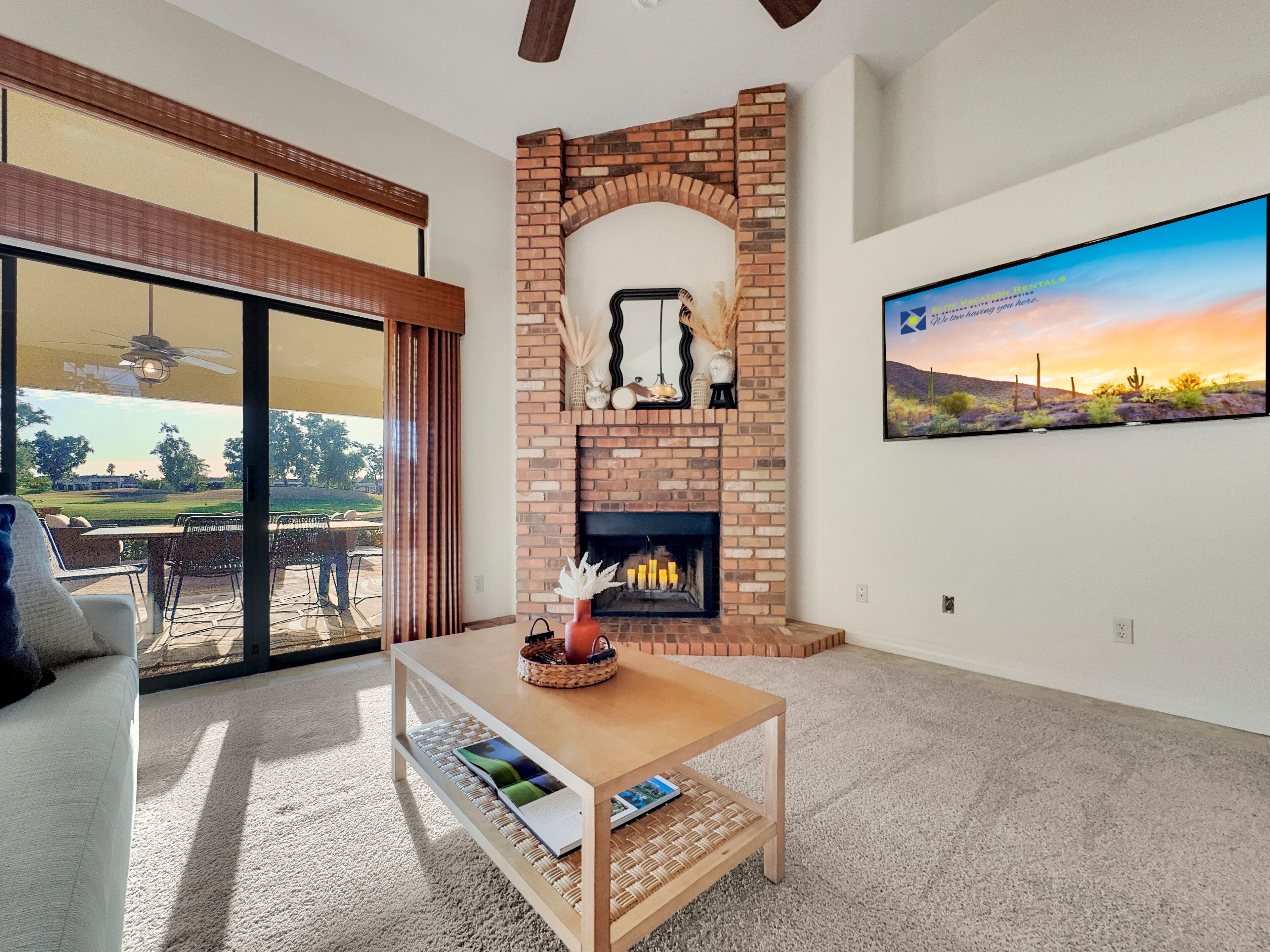 A living room with a brick fireplace, a television mounted on the wall, and a view of a golf course through large sliding glass doors.