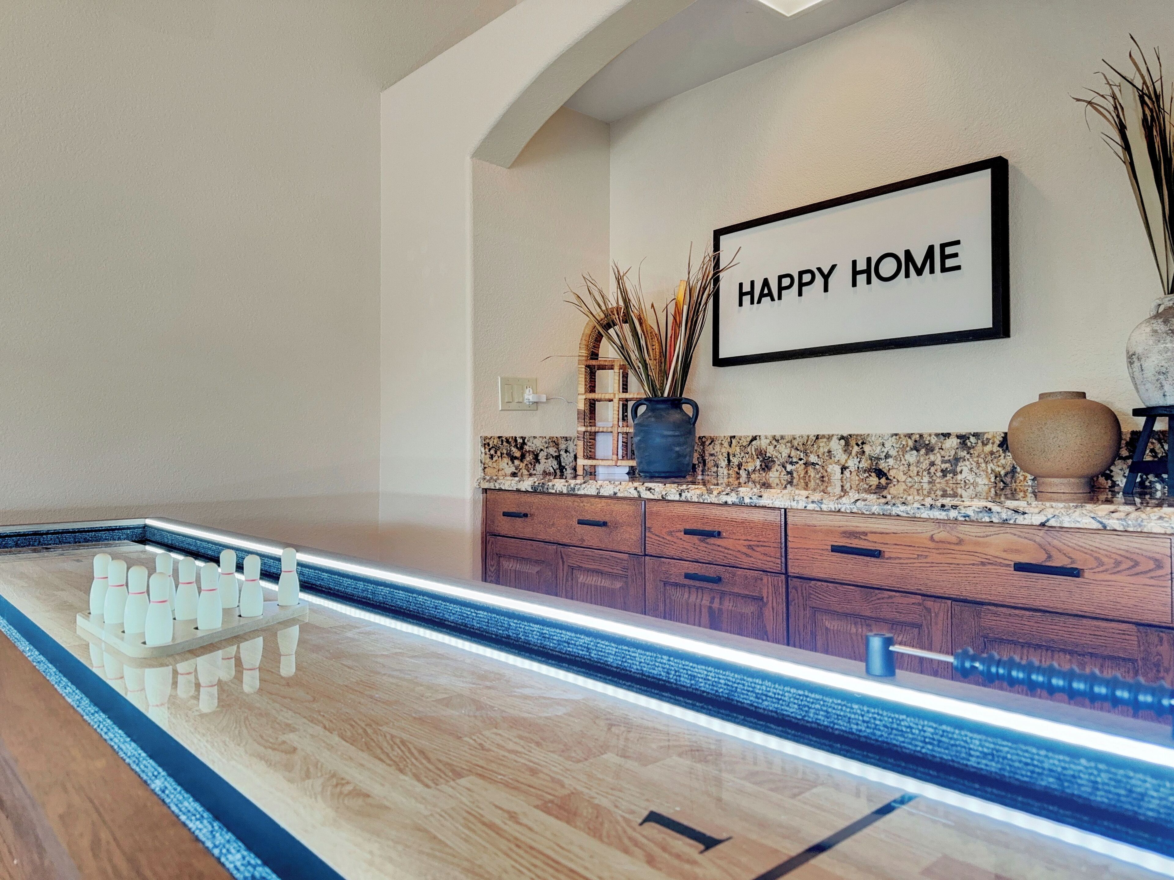 A modern kitchen with a bowling alley setup, wooden cabinets, granite countertop, and a 'HAPPY HOME' sign on the wall.