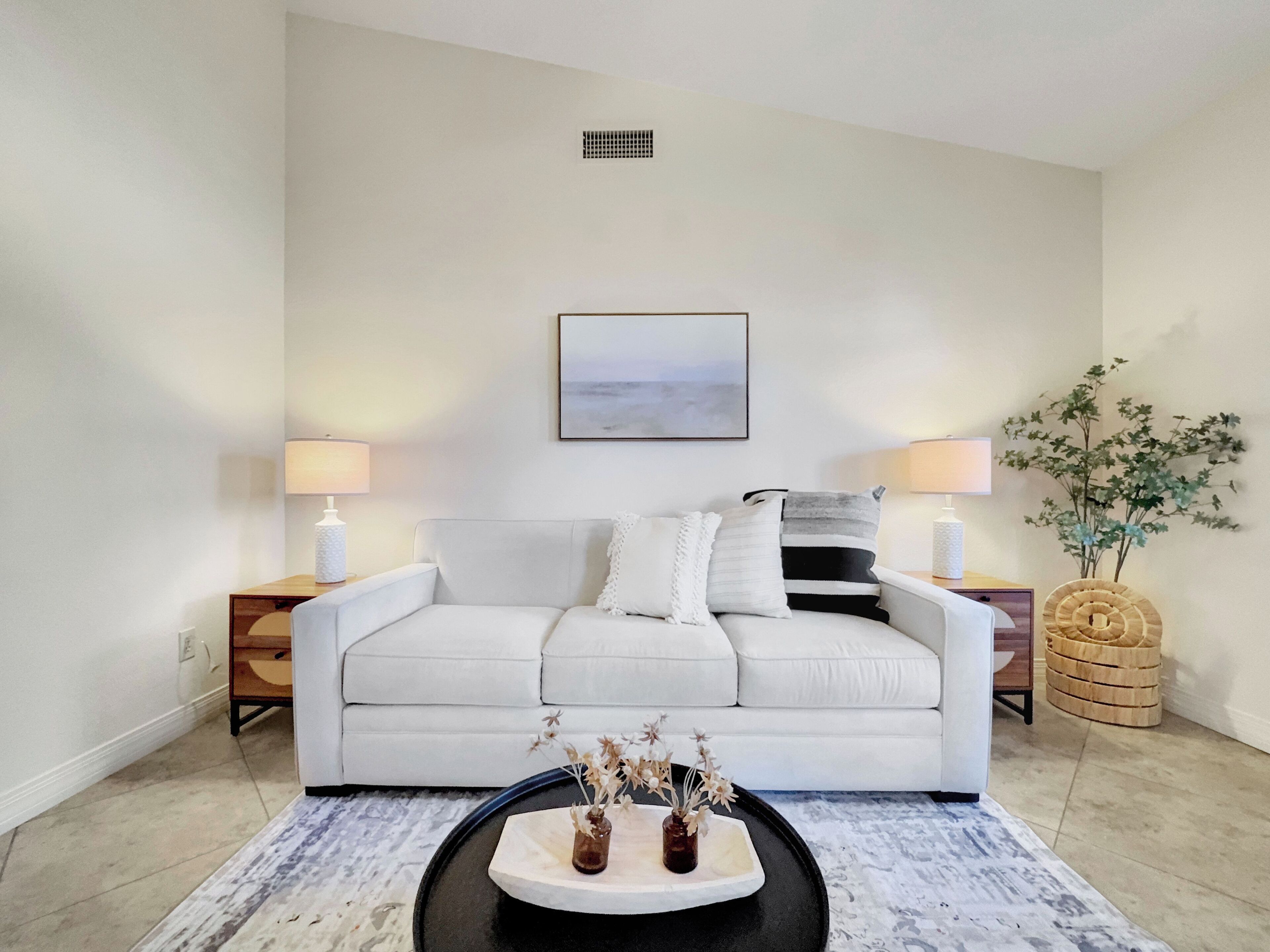 A modern living room with a white sofa, two side tables with lamps, a coffee table with decorative items, a framed picture on the wall, and a potted plant.