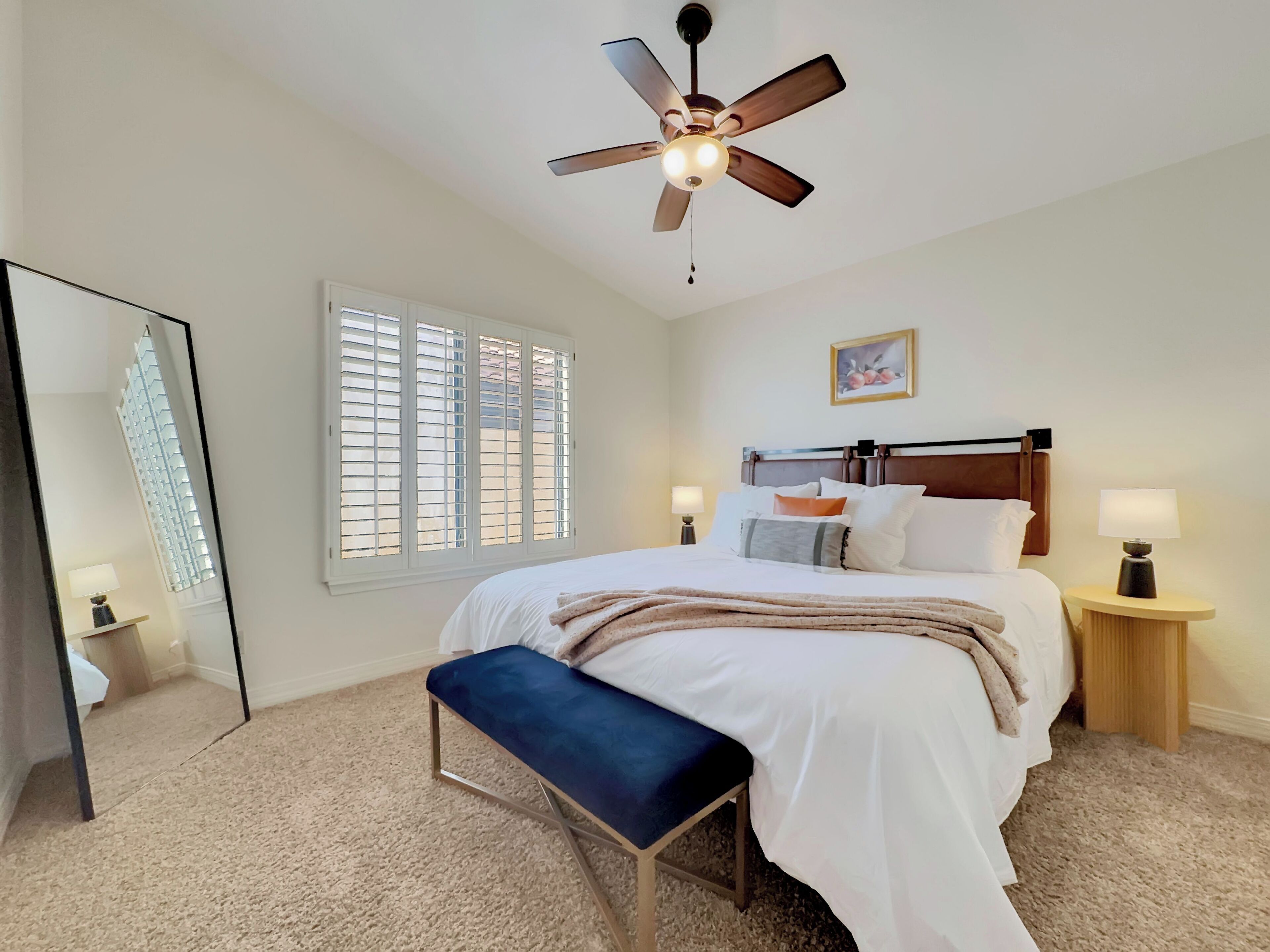 Quarto de hotel com cama, um banquinho, um ventilador de teto, janela com persianas, mesa de cabeceira com luminária e um espelho.
