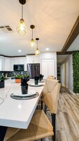 Una cocina moderna con armarios blancos, un refrigerador de acero inoxidable y un comedor con una isla blanca y sillas de mimbre.