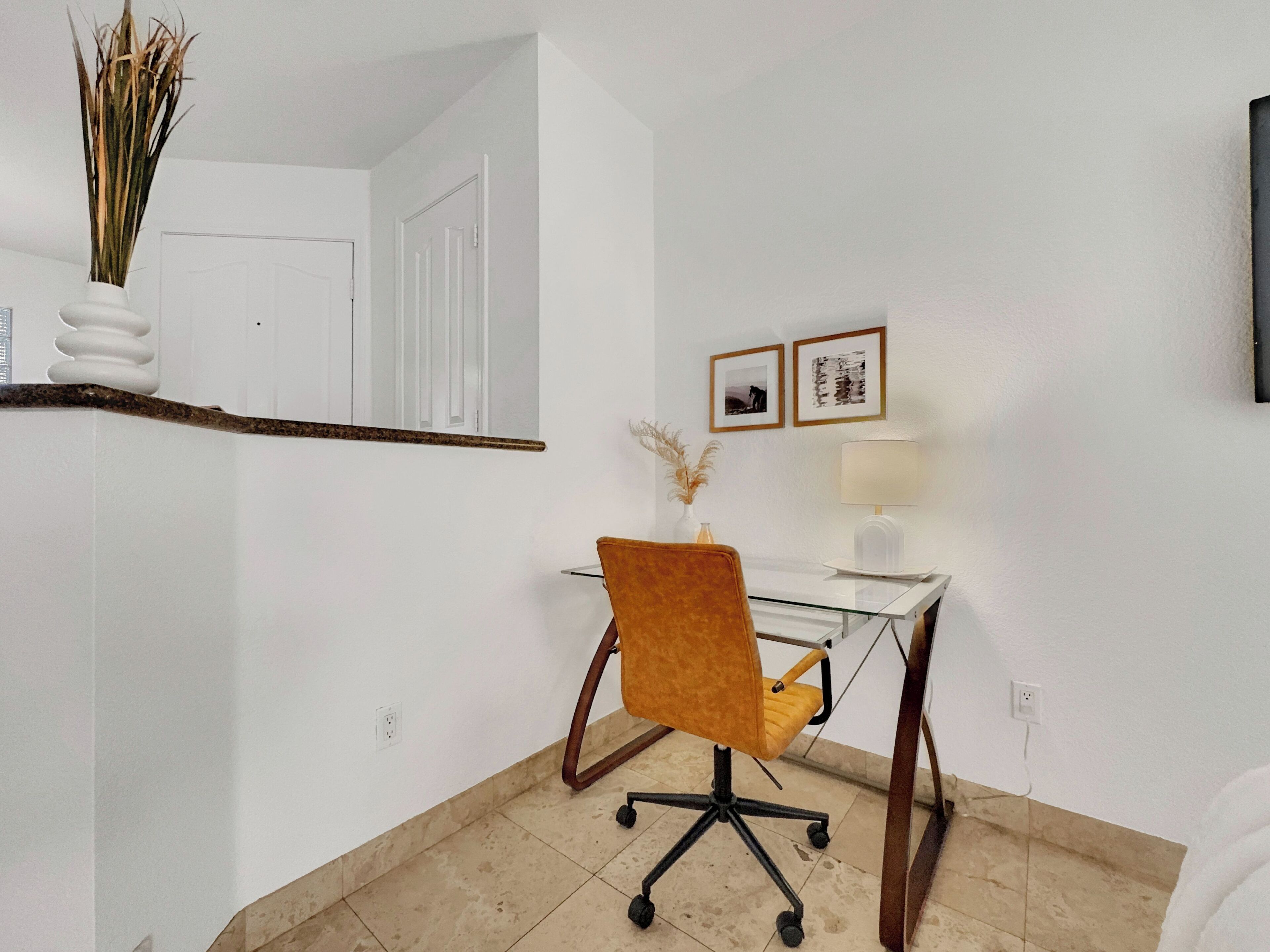 Un petit bureau avec un bureau en verre, une seule chaise orange, un vase blanc contenant des plantes séchées, une lampe blanche et deux tableaux encadrés accrochés au mur.