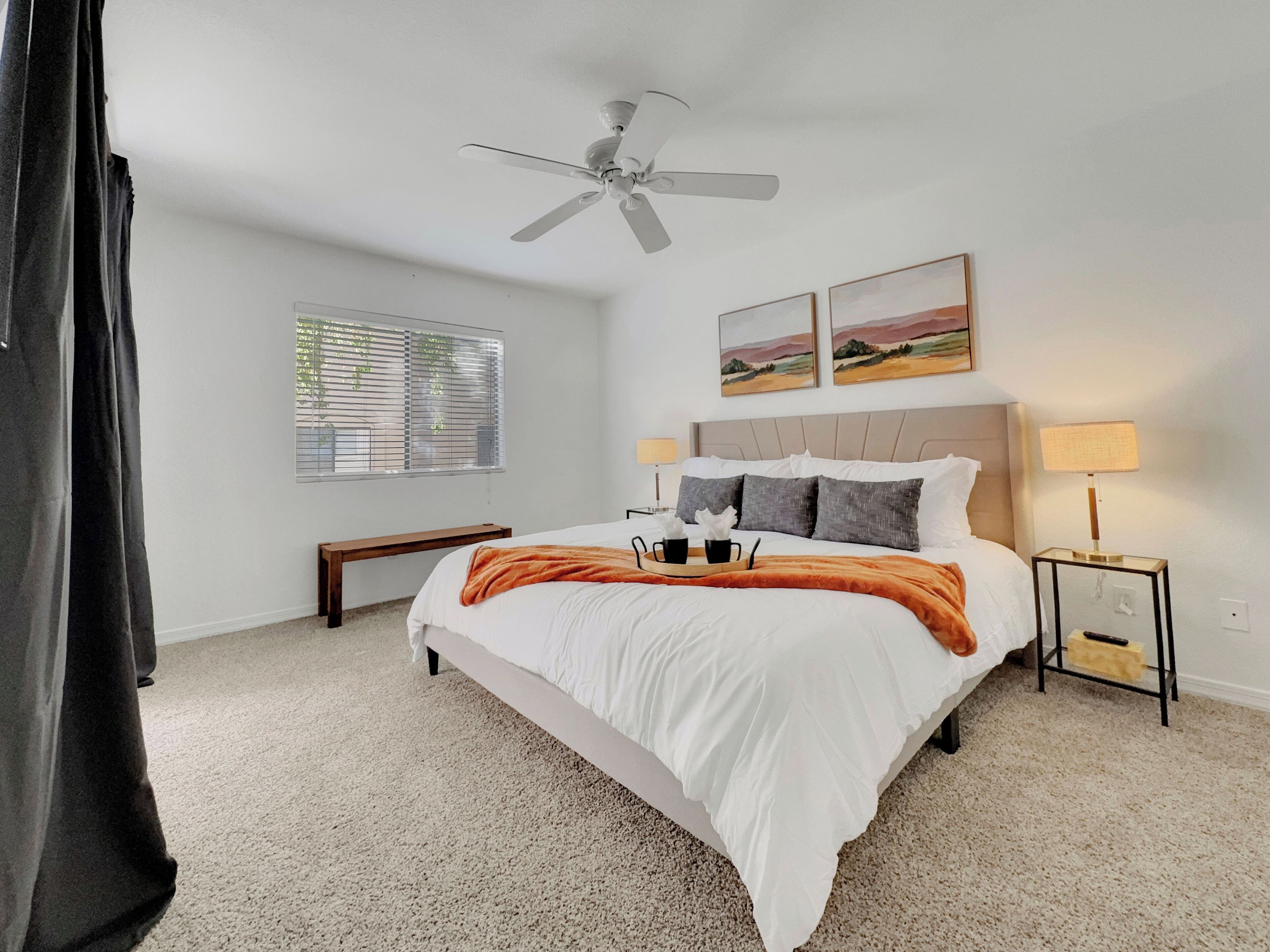Quarto com cama, ventoinha no teto, mesa de cabeceira com abajur, janela com persianas e duas pinturas de paisagens na parede.